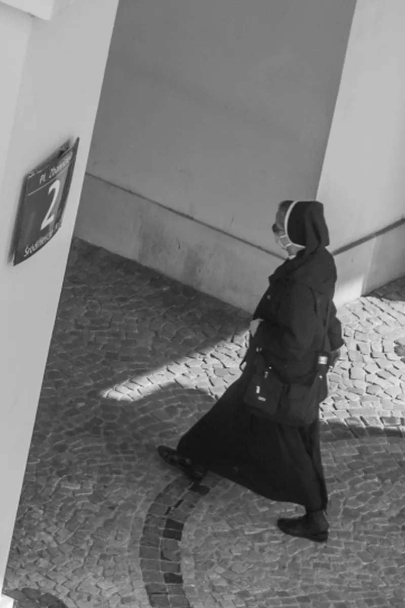 Masked nun walking alone across a curved cobblestone path